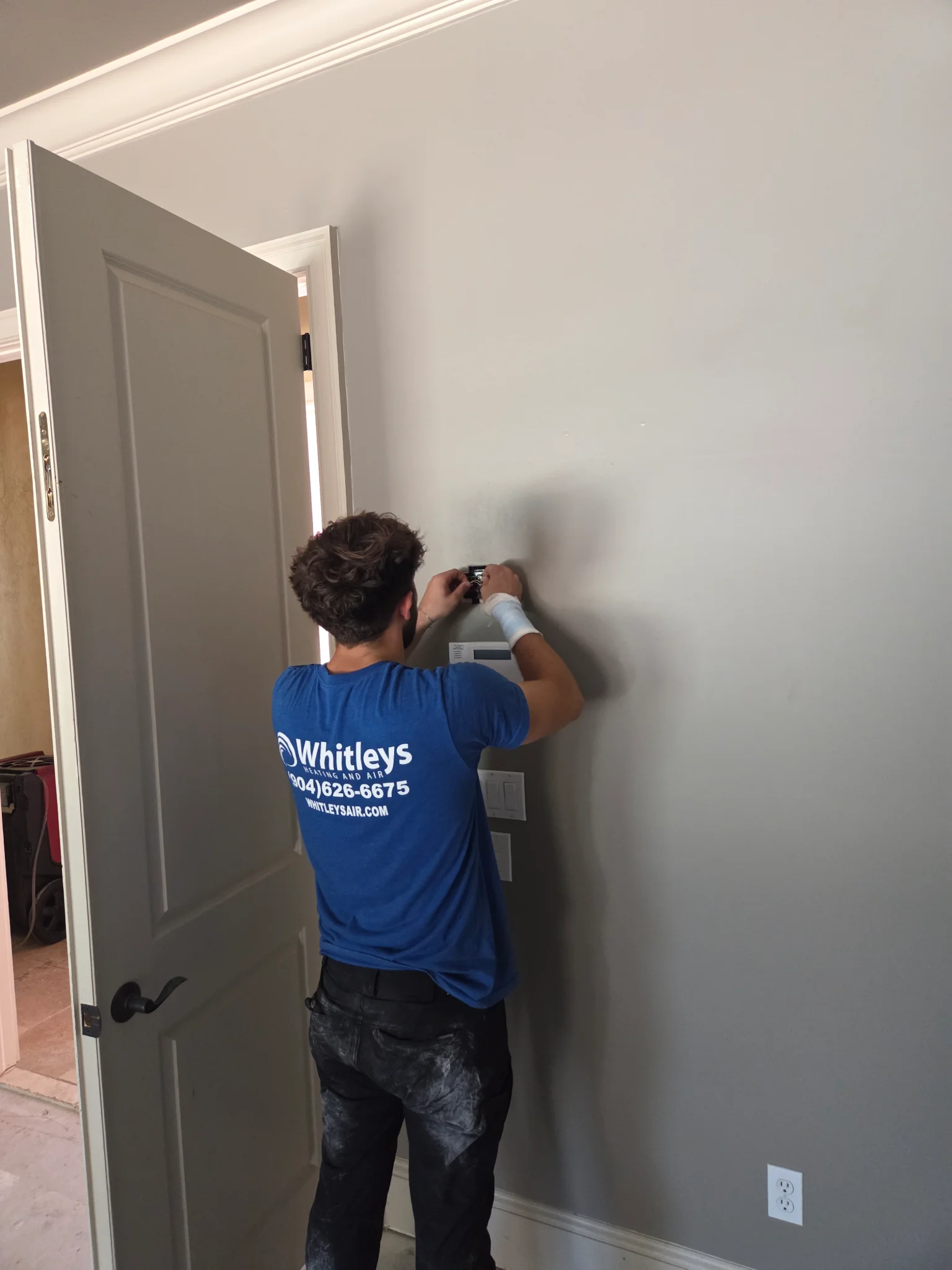 A man wearing a blue t-shirt and black pants is installing a light switch on a light gray wall. The  