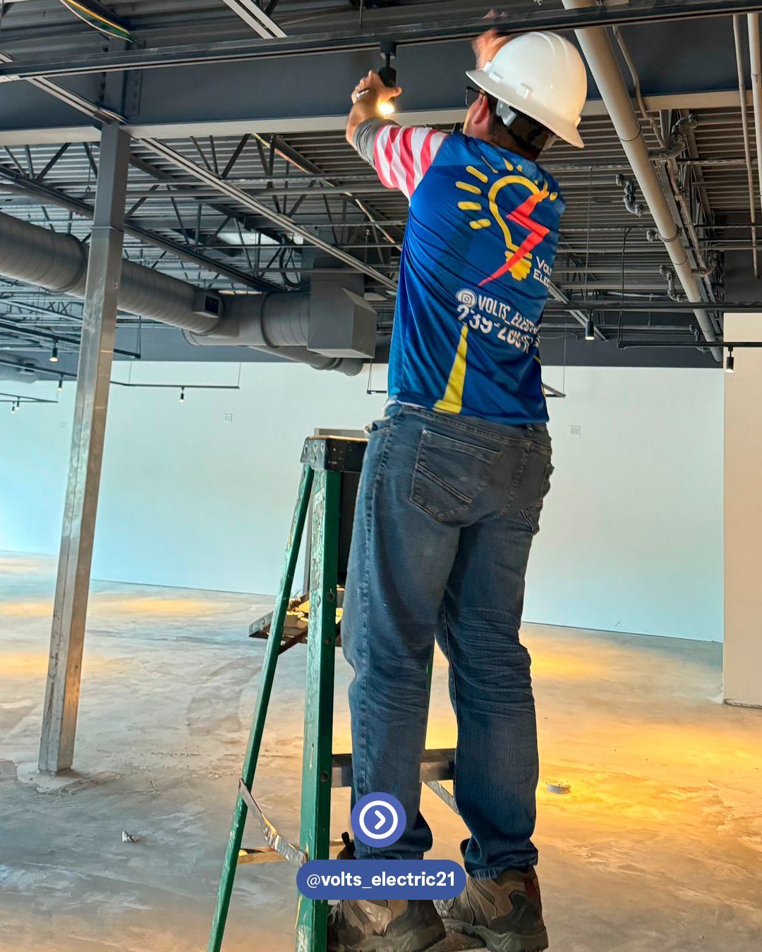 Electrician standing on a ladder installing or inspecting ceiling electrical wiring and lighting inside a commercial building with exposed ductwork and metal framing, wearing safety gear and a Volts Electric shirt.
