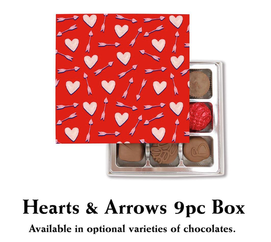 A Valentine's Day-themed table featuring Florence's Exquisite Chocolates in Rexburg, Idaho. At the center is a beautifully packaged red box of 9 chocolates adorned with heart and arrow designs, labeled "Hearts & Arrows 9pc Box." The box contains an assorted selection of chocolates, including Milk Chocolate, Dark Chocolate, and varieties like Assorted Chocolates, Nuts, and Caramels, arranged neatly in a silver tray. The table is adorned with romantic touches such as heart-themed plates, decorative candy bowls, festive napkins, and red roses. This inviting setup highlights Florence's chocolates as the perfect gift for Valentine’s Day.