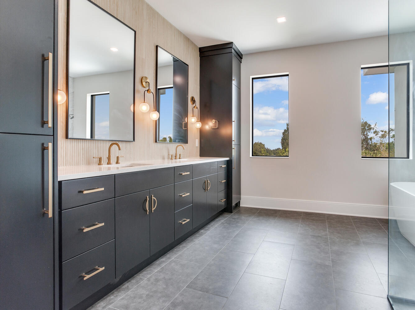 Spacious primary bathroom with double vanity, dark custom cabinetry, large mirrors, freestanding tub area, and contemporary luxury finishes.