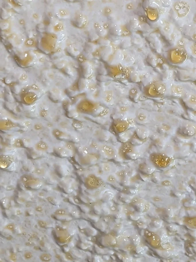A close-up, macro photograph captures a textured surface, possibly a food item like frosting or dough, with glistening droplets of a golden liquid, likely honey or syrup, scattered across its uneven landscape. The lighting highlights the sheen on the droplets and the peaks and valleys of the textured surface, creating a rich, appetizing visual.