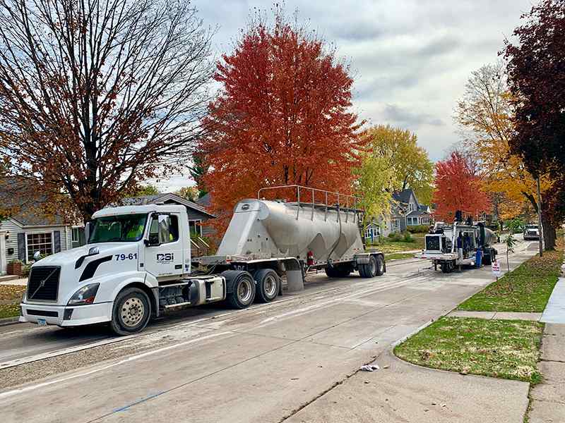 Street-level view along a quiet residential road lined with vibrant red and yellow autumn trees. A white DSI support truck towing a specialized hopper/tanker trailer is parked near the curb, with a compact grinding unit working further down the street.