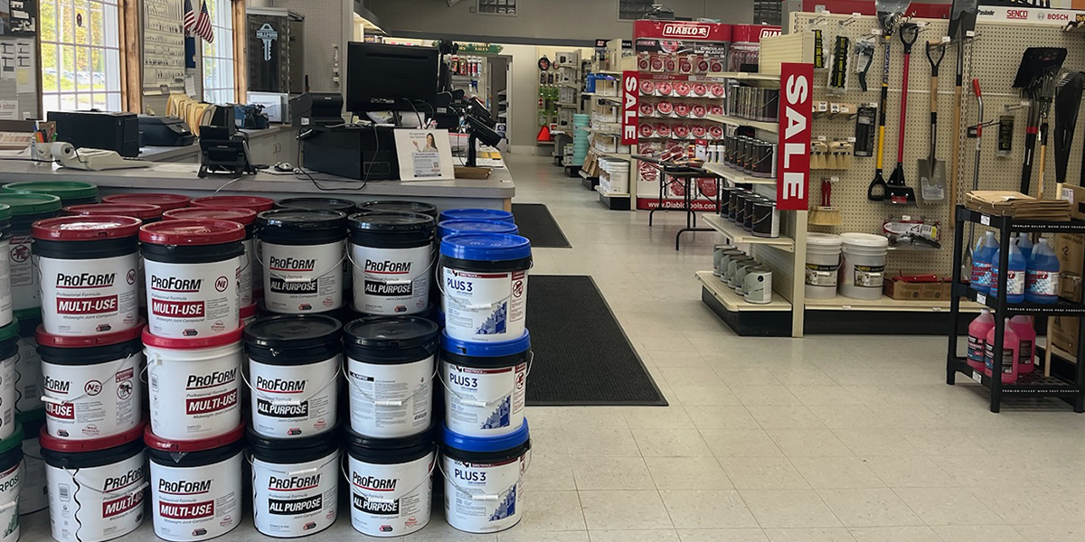 A retail counter and interior view of Builders FirstSource, displaying stacks of ProForm Multi-Use and All-Purpose compound buckets alongside shelves stocked with tools, paints, and construction essentials. The layout highlights a clean and well-organized shopping experience for contractors and DIY customers.