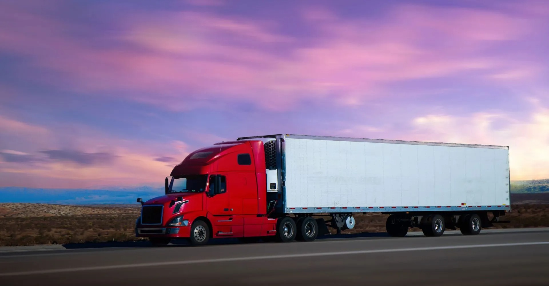 Red semi truck pulling a dry van trailer across an open roadway. Long-distance freight transportation supporting commercial shipping, distribution networks, and reliable cargo delivery services.