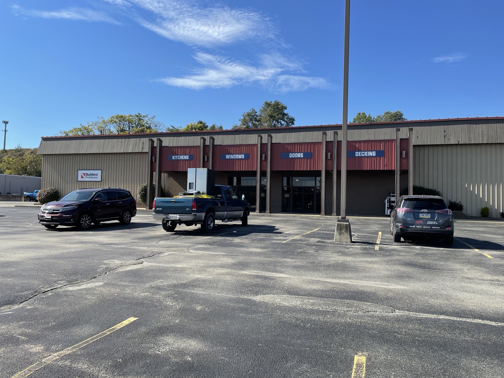 Builders FirstSource showroom entrance displaying product categories including kitchens, windows, doors, and decking, with customer parking and vehicles visible in front of the building.