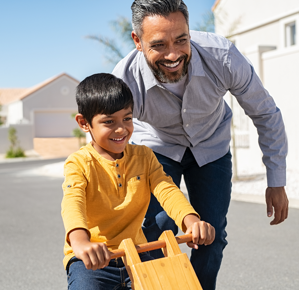 A father pushing his young son down the street on his tricycle.