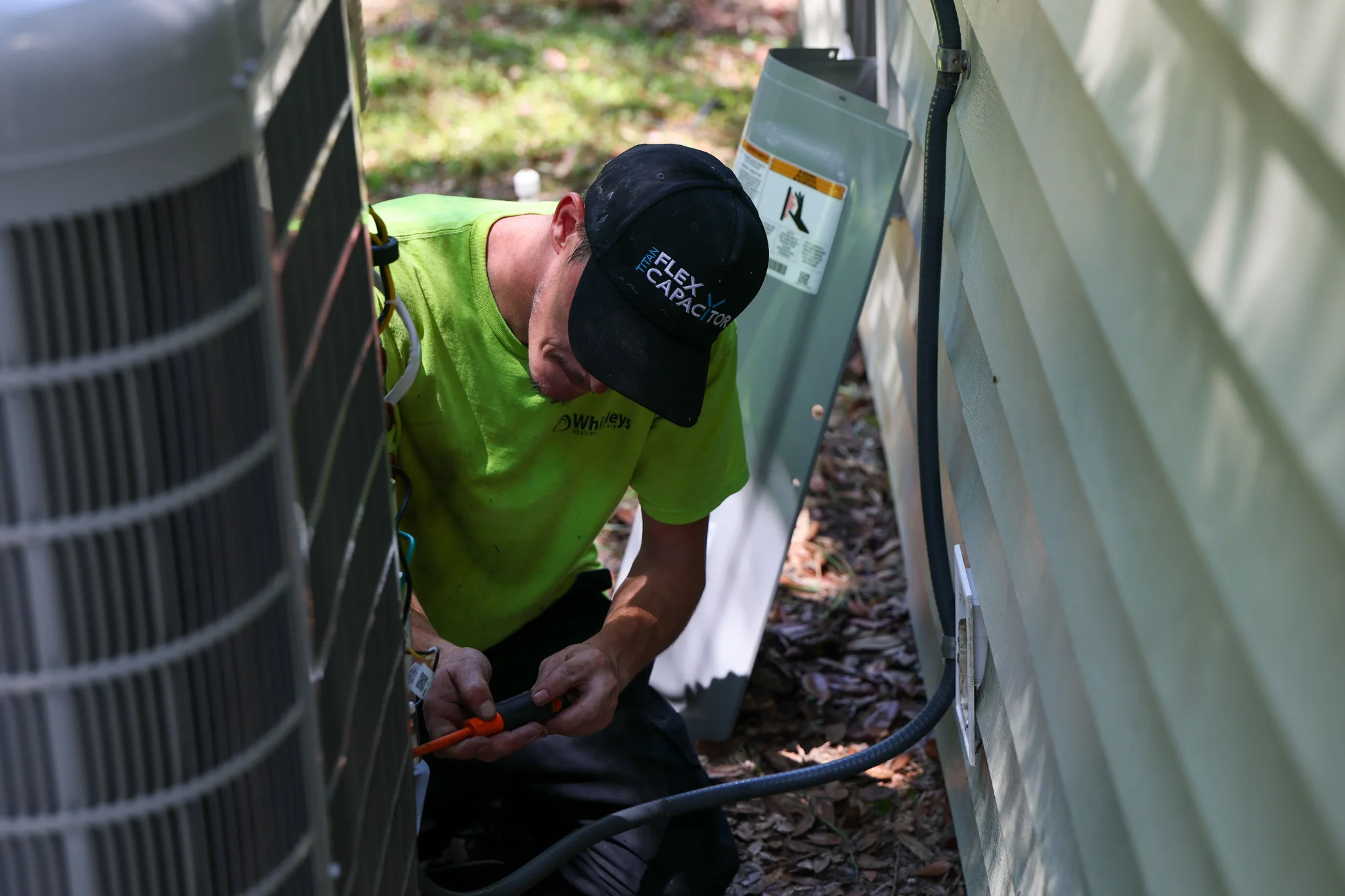 A man in a bright green t-shirt and black baseball cap is kneeling down and working on an air condit 