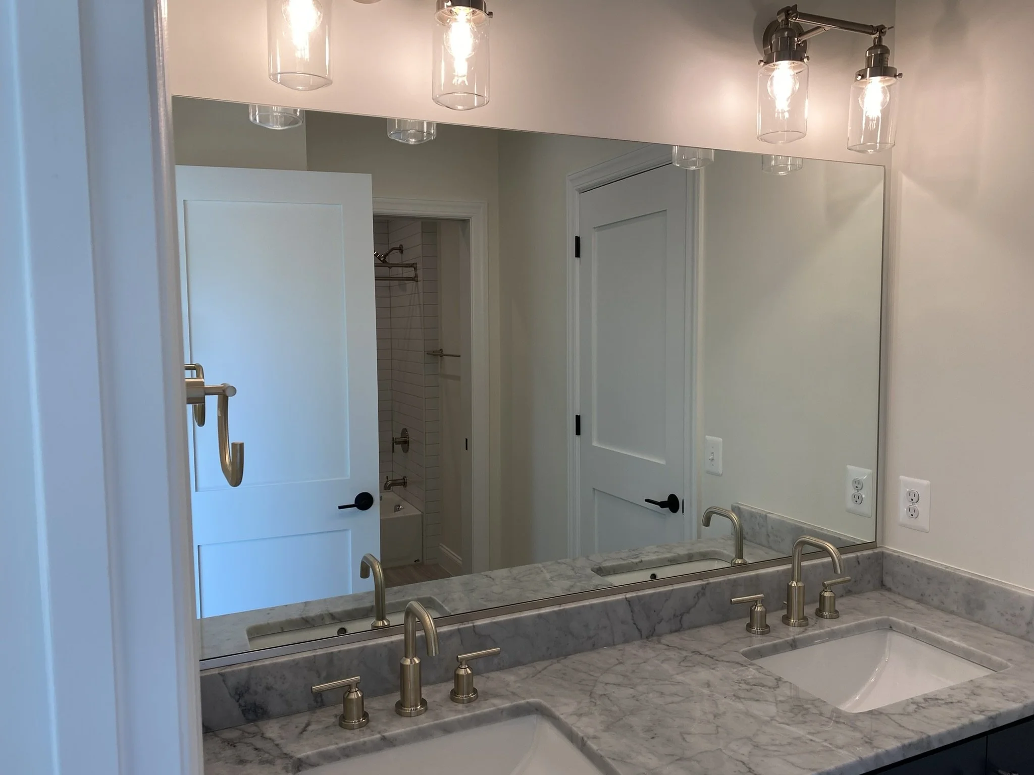 A well-lit bathroom vanity area with a large mirror reflecting doors and a shower. The vanity features two sinks with modern brushed nickel faucets, set into a marble countertop. Above the mirror, a light fixture with three glass-shaded bulbs illuminates the space. Electrical outlets are visible on the wall.