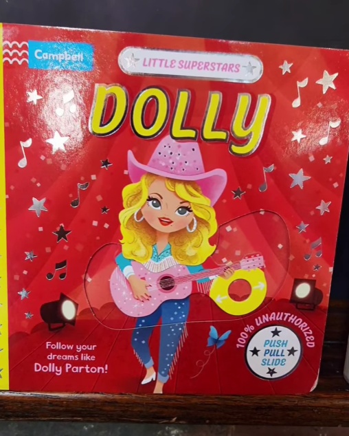 Dressed in a Dolly Parton shirt, a staff member at Noggin Factory in Dover, NH, showcases the interactive board book Little Superstars: Dolly, highlighting its fun wig page and sliding features.