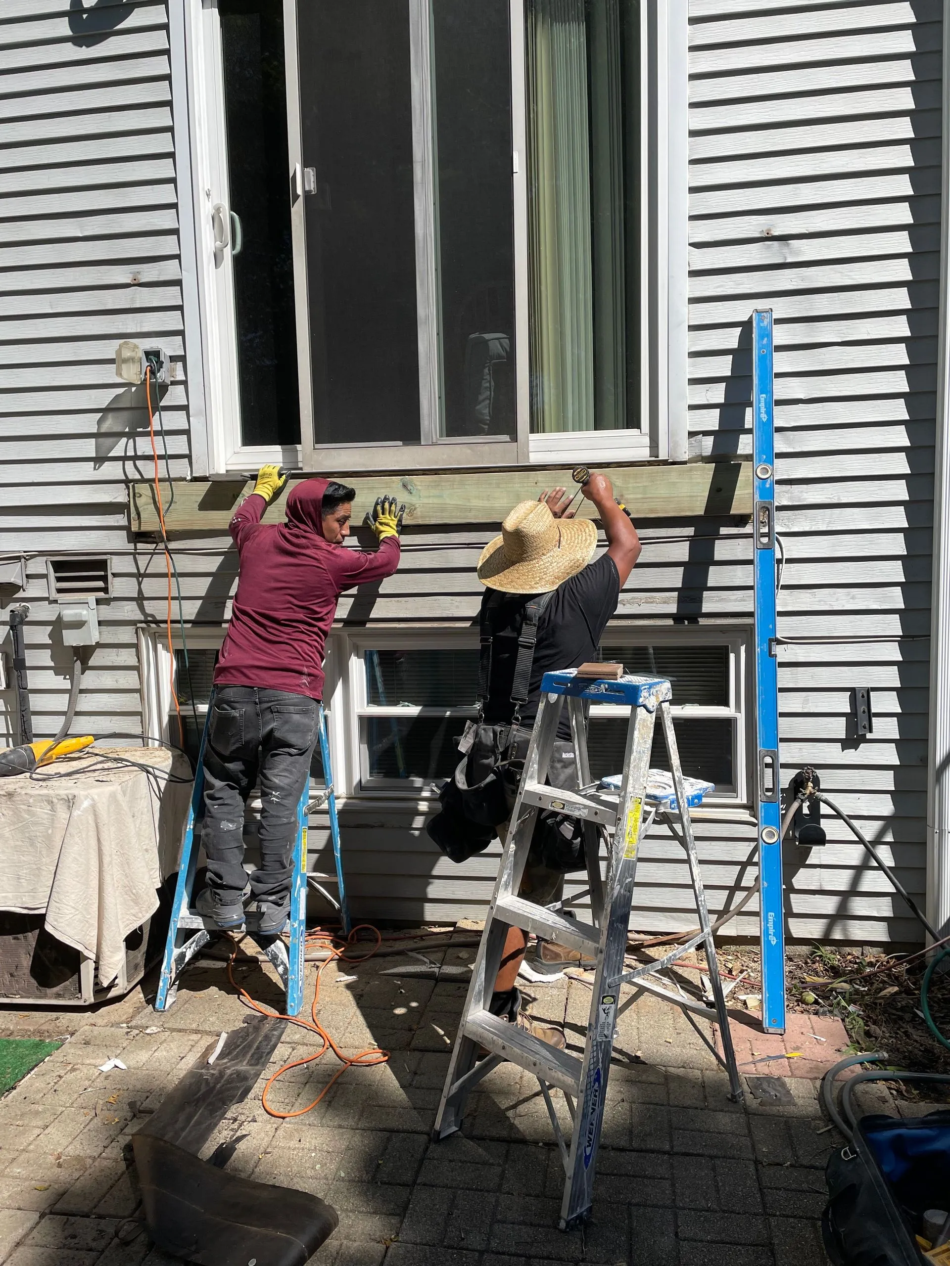 Contractors install a deck ledger board beneath a sliding patio door. Ladders, tools, and safety equipment highlight professional preparation for a secure and properly supported deck structure.