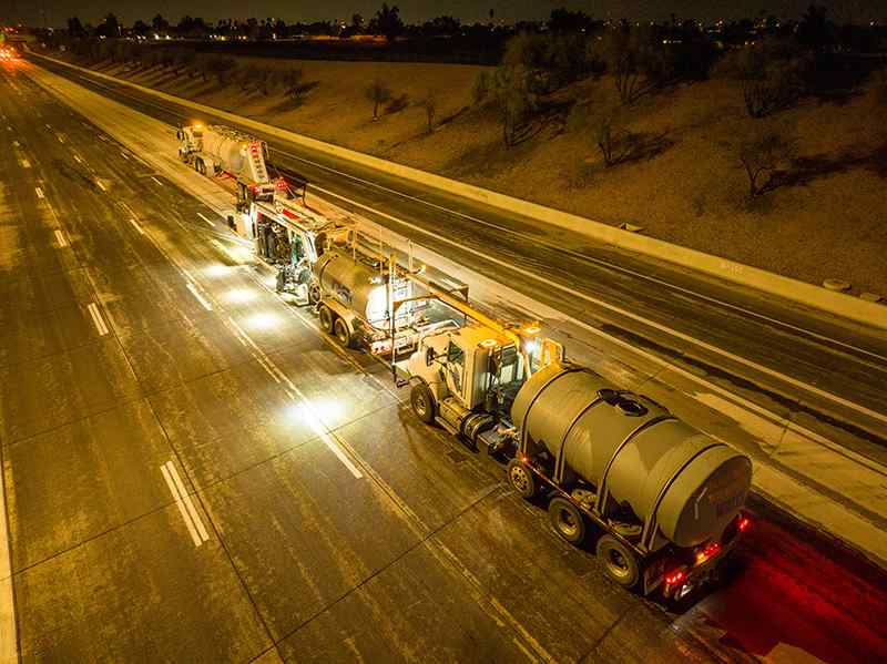 High-angle night photograph of a night-shift grinding operation on a multi-lane highway. Illuminated by bright work lights, a full DSI grinding train—including a tanker truck and grinding rig—operates along the lane with long-exposure traffic lights glowing in the distance.