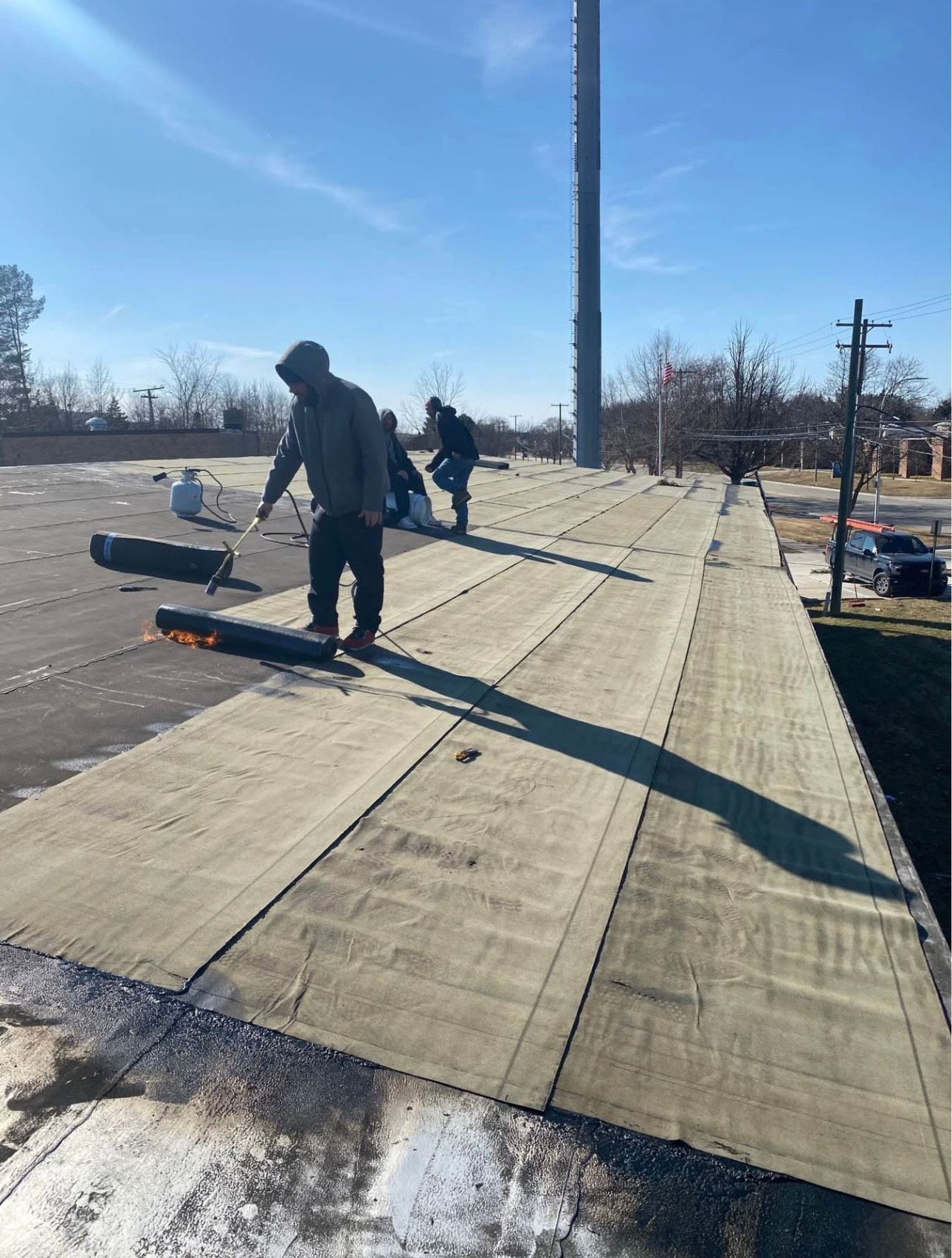 On a sunny day, three workers install membrane on a long, flat roof. The foreground worker uses a propane torch, with the flame visible, to apply a roll of roofing material.