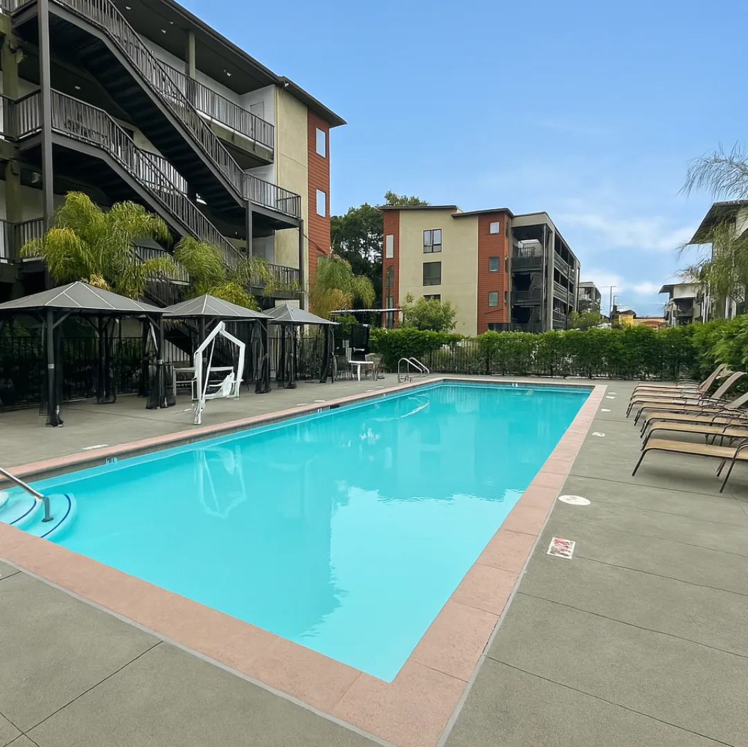A swimming pool surrounded by lounge chairs and umbrellas in front of apartment buildings.