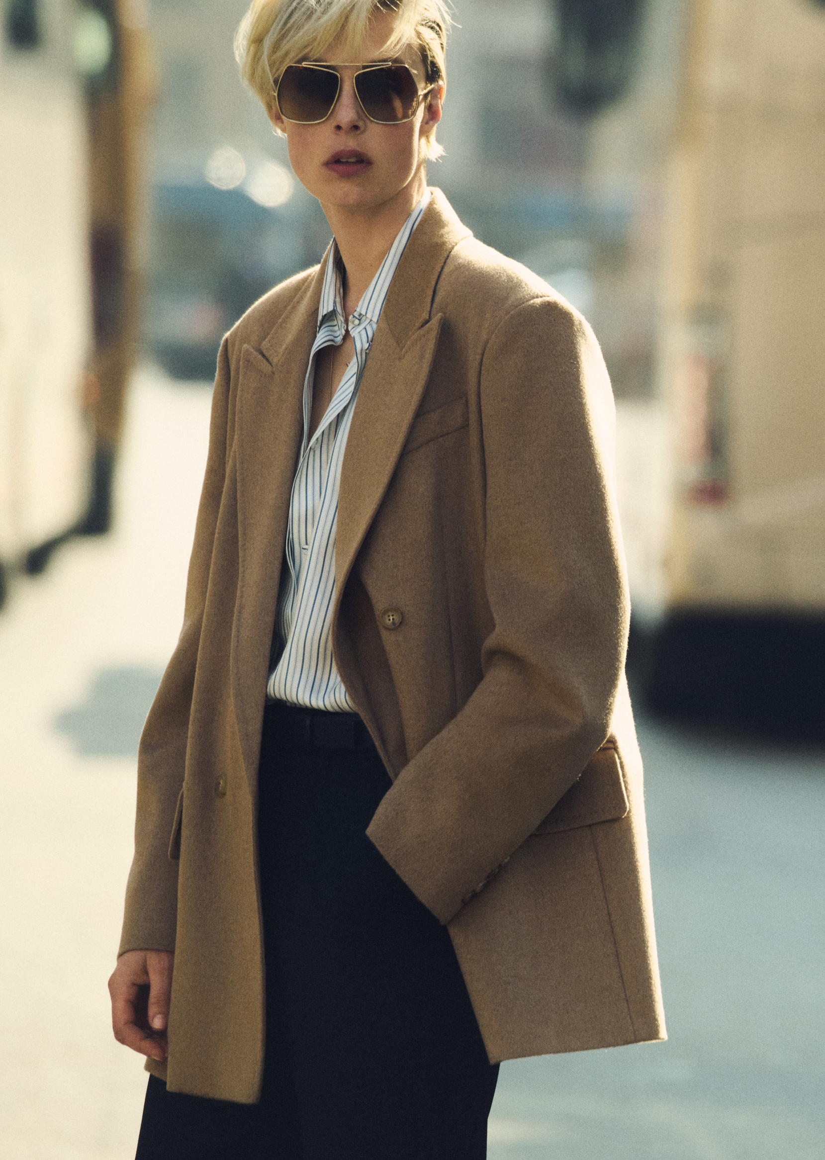 Модель в двубортном блейзере Olimpia Jacket от Max Mara цвета верблюжьей шерсти, темных очках и рубашке на фоне городского пейзажа.