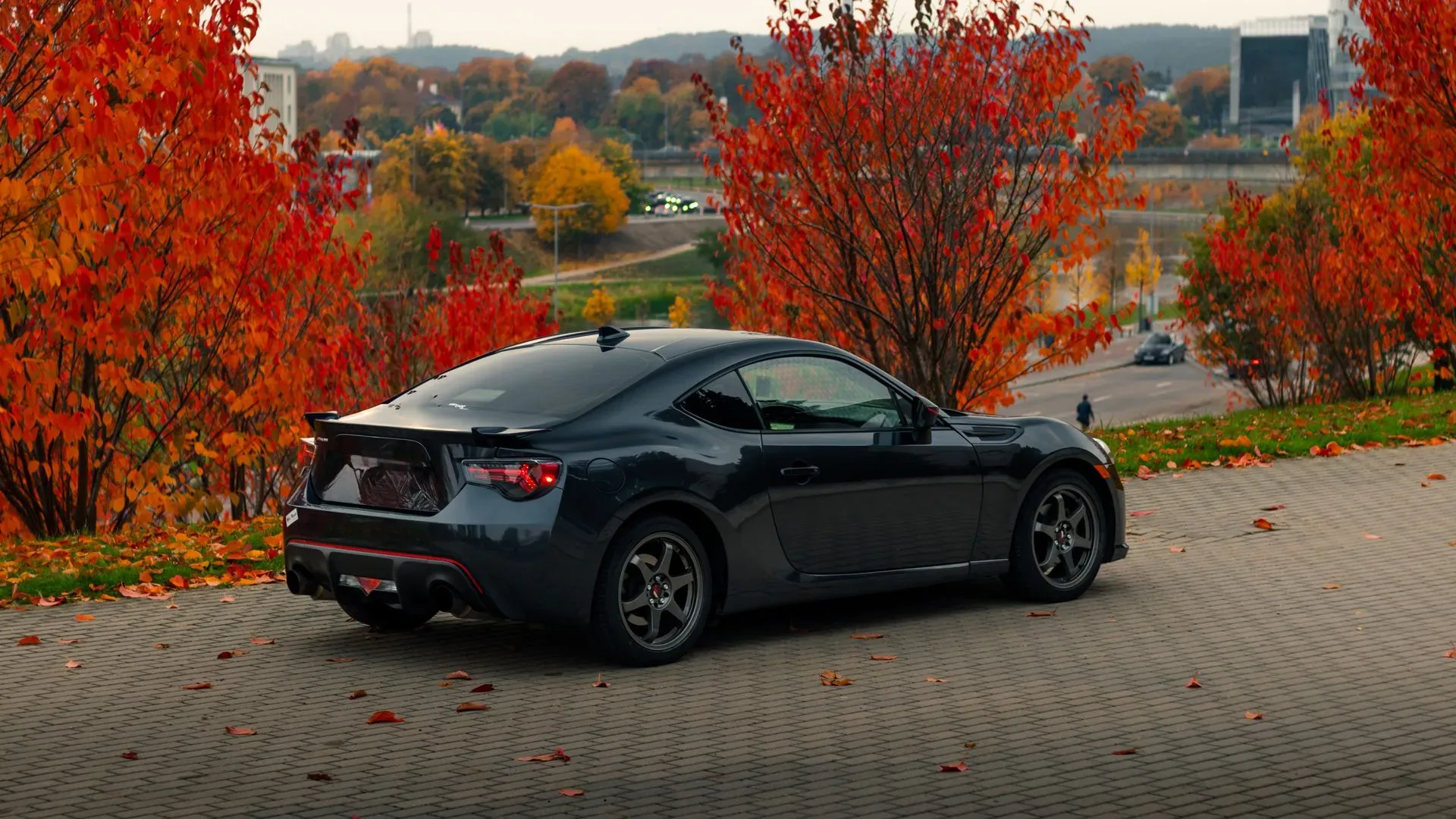 A dark grey Subaru sports car is parked on a cobblestone driveway, surrounded by vibrant autumn foliage. The leaves on the trees are a mix of bright red and orange, creating a striking contrast with the car and the overcast sky. Fallen leaves are scattered across the ground. In the background, a city skyline and a body of water can be seen, along with other vehicles on a distant road.