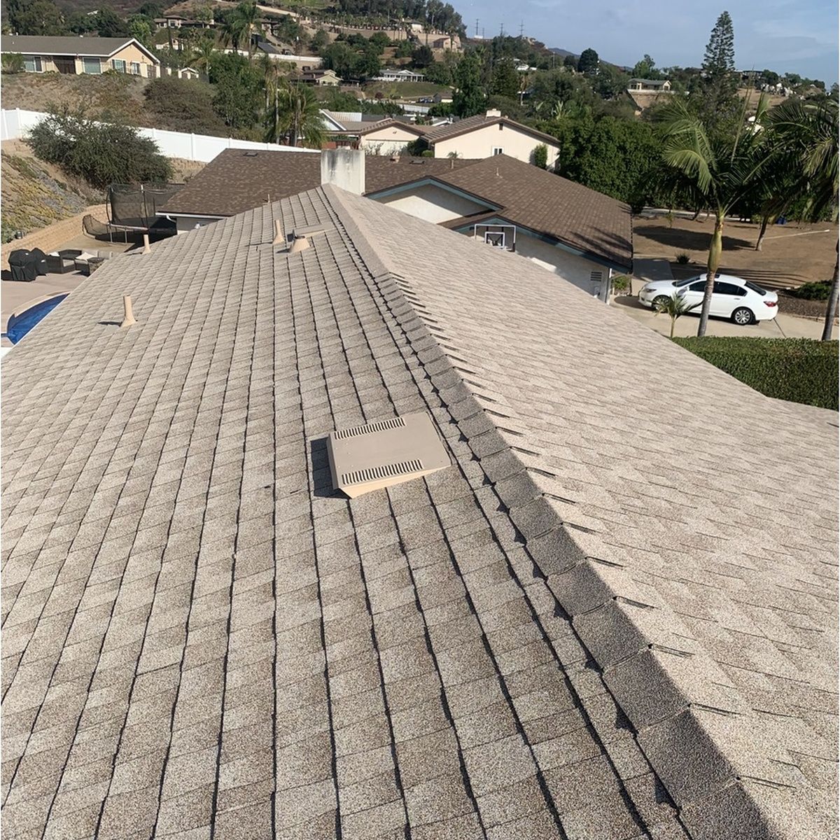 Overhead view of a large residential roof featuring tan architectural shingles, central roof vents, and pipe boots, overlooking a scenic hillside neighborhood with palm trees.