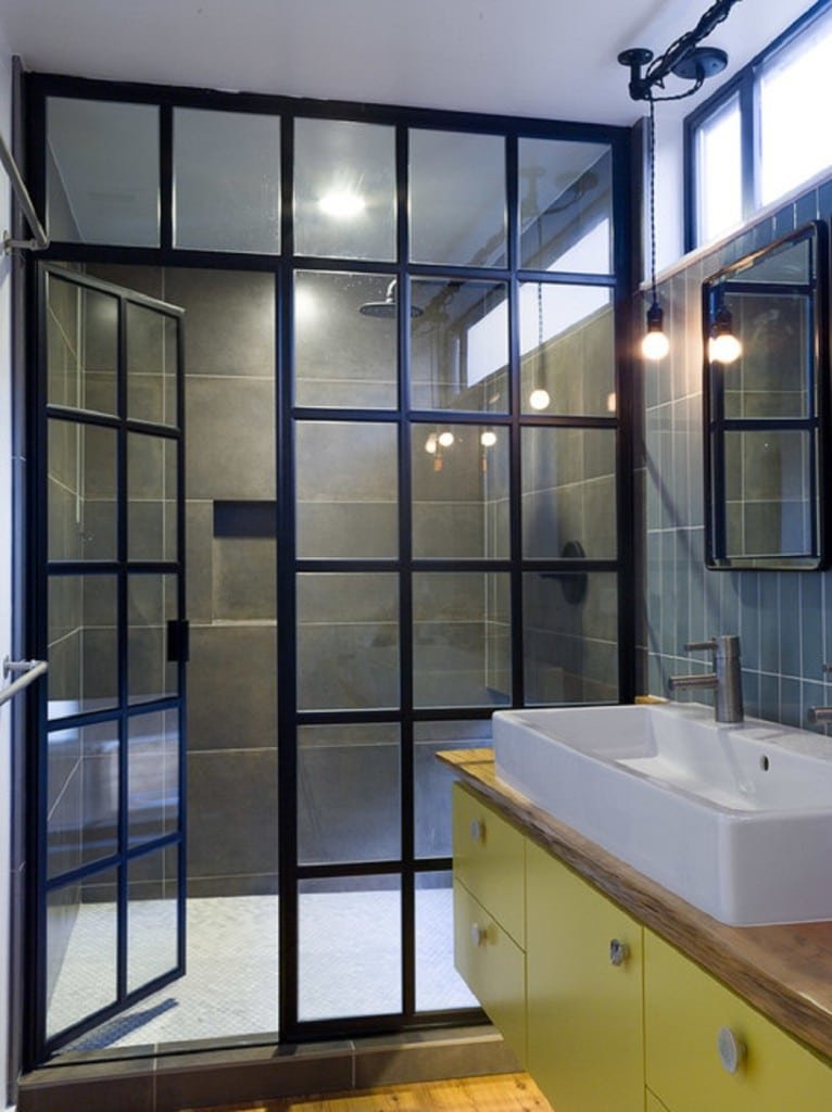 A modern bathroom featuring a walk-in shower with a black metal frame and glass doors, a white rectangular sink mounted on a bright yellow vanity, and a large mirror on a tiled wall.