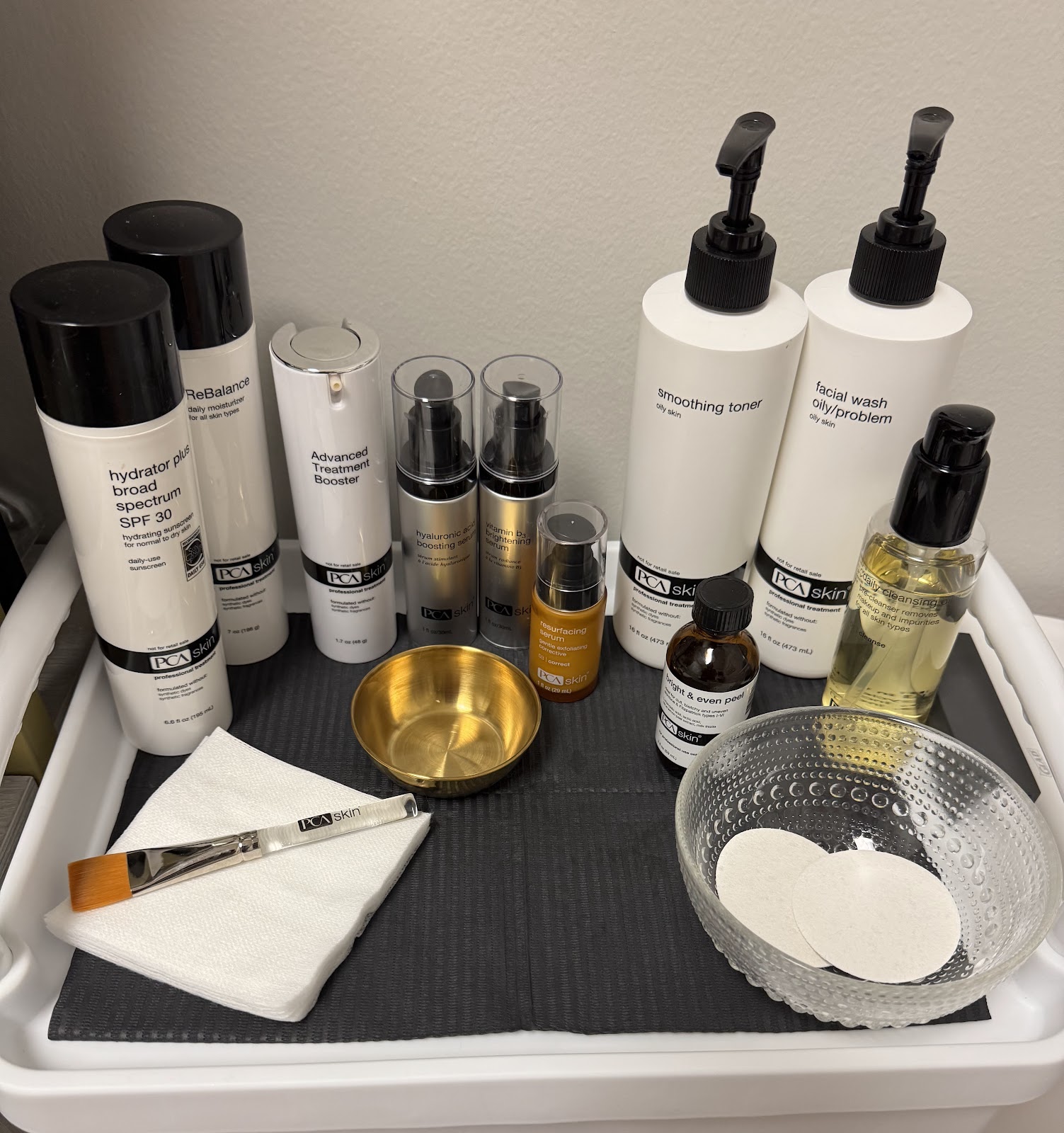 A collection of PCA SKIN professional skincare products are neatly arranged on a white tray with a black mat. The products include a broad-spectrum SPF 30 hydrator, a daily moisturizer, an advanced treatment booster, a hyaluronic acid boosting serum, a vitamin B3 brightening serum, a resurfacing serum, a smoothing toner, a facial wash for oily/problem skin, a brightening & even peel, and a daily cleansing oil. A small brass bowl, a glass bowl with cotton pads, a facial brush, and a stack of white paper napkins are also present.