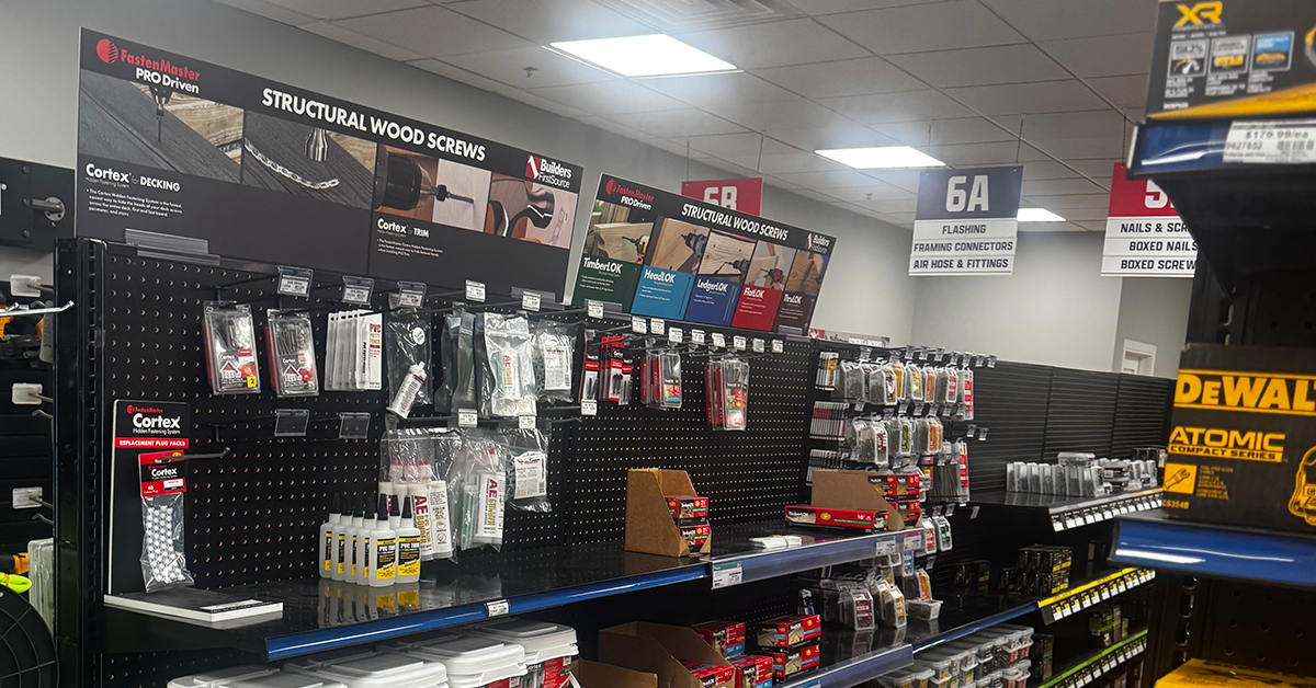 Wide view of a Builders FirstSource store aisle with black pegboard displays of structural wood screws, decking fasteners, adhesives, hardware, and white buckets of Cortex products. Overhead signs identify sections for structural wood screws, flashing, framing connectors, air hoses, fittings, nails, and screws. Yellow DEWALT boxes appear on shelving at the right.