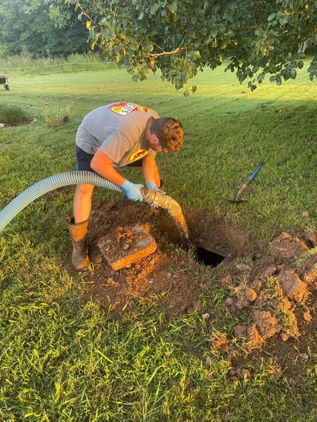 A service technician in a grey shirt and boots pumps a residential septic tank using a heavy-duty hose. The work is performed in a grassy yard near a small excavation.