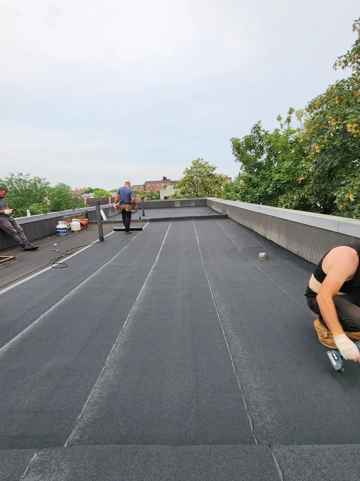 Three workers are engaged in installing a dark membrane on a large flat roof. One kneels working near the edge, another stands centrally, and a third rests near supplies.