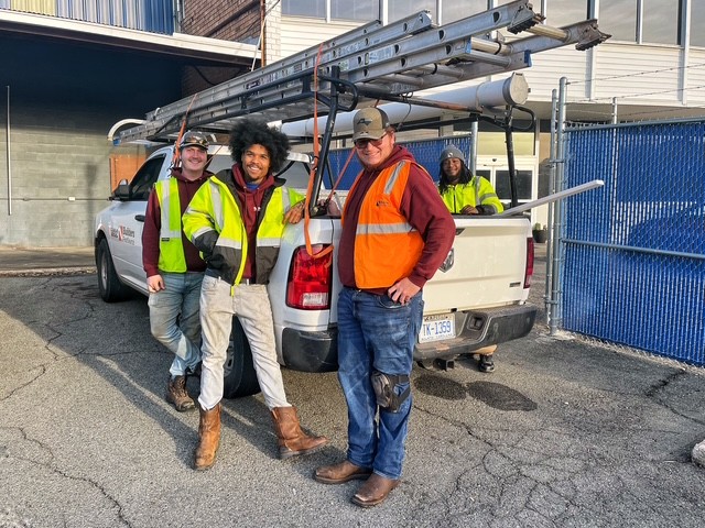 Four workers wearing safety vests and work boots stand beside a white pickup truck parked in an industrial area. A long ladder is secured to a rack on top of the truck, and metal pipes extend from the truck bed. Buildings, fencing, and a paved lot are visible in the background.