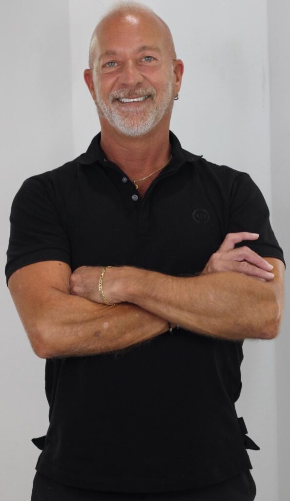 A medium shot portrait of a bald, middle-aged man with a gray beard and mustache. He is wearing a black polo shirt and has his arms crossed. He is smiling and looking at the camera. There is a gold chain around his neck and a gold bracelet on his left wrist. His skin has some patches of discoloration.