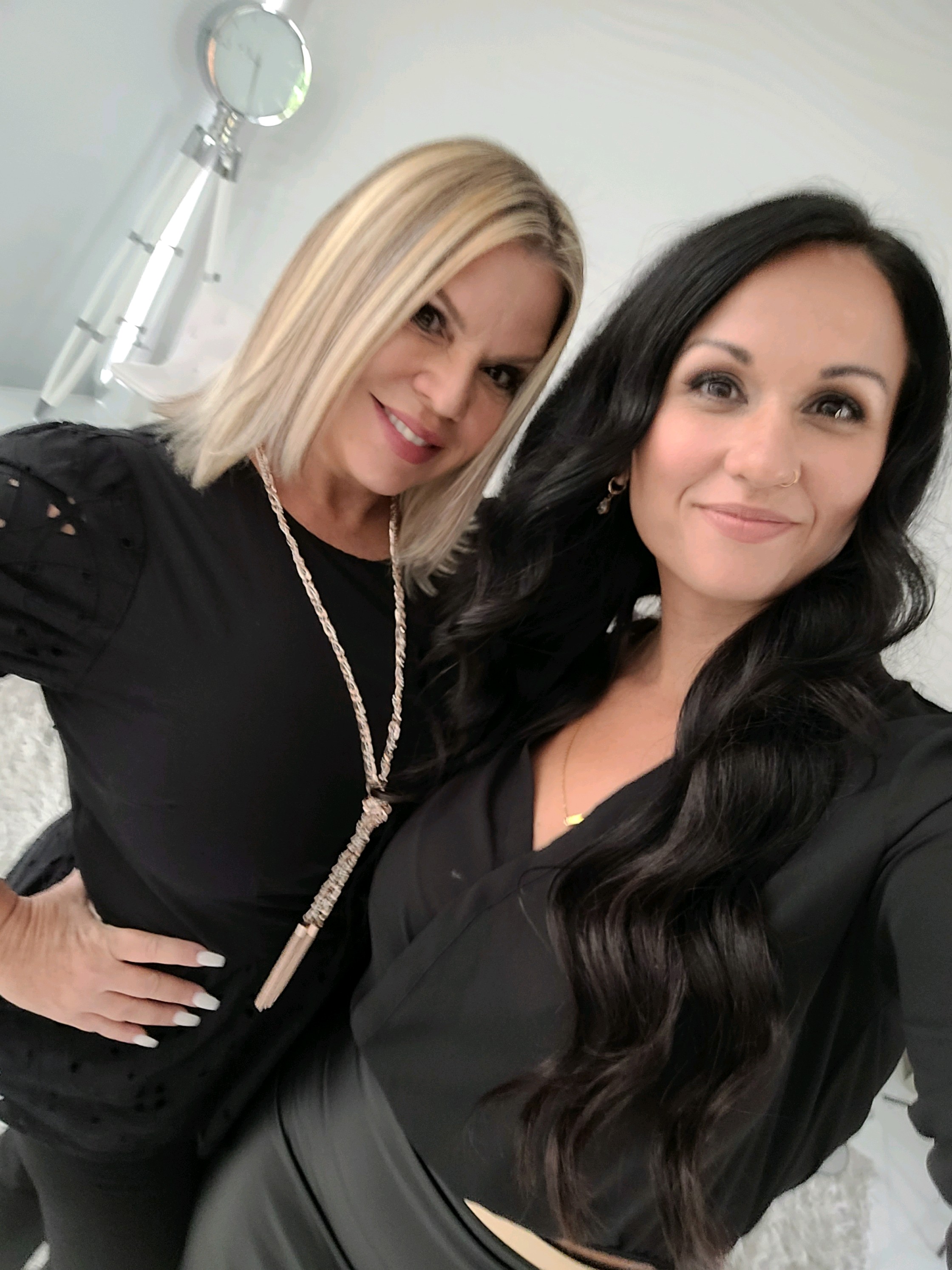 Two women in black clothing are posing for a selfie in a brightly lit room. The woman on the left has blonde hair and is wearing a black top with eyelet details on the sleeves and a long, chunky necklace. The woman on the right has long, dark hair and is wearing a black top with a gold necklace. Both women are smiling at the camera. The background appears to be a well-lit interior with a hint of a modern lamp or fixture visible on the left.