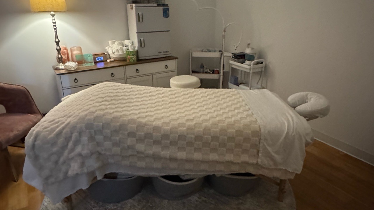 A massage or spa room is shown, featuring a massage table covered with a checkerboard patterned blanket and white linens. There is a pink chair to the left, a dresser with various items on top including a lamp, decorative items, and a digital clock displaying '05:26'. A towel warmer or cabinet is visible in the background, along with shelves holding spa supplies and equipment. The room appears to be set up for therapeutic or relaxation treatments.