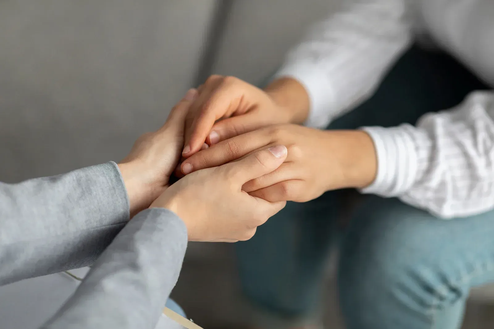 The image shows two individuals holding hands in a close-up shot. One person wears a light gray long-sleeved shirt, while the other wears a white long-sleeved shirt and blue jeans. Their hands are intertwined, suggesting comfort, support, or empathy.
