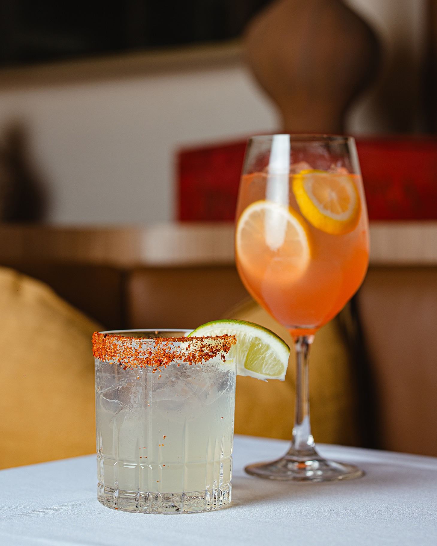 Two refreshing cocktails: a rocks glass with a Tajin rim and lime garnish next to a tall wine glass filled with an orange-hued drink and lemon slices.