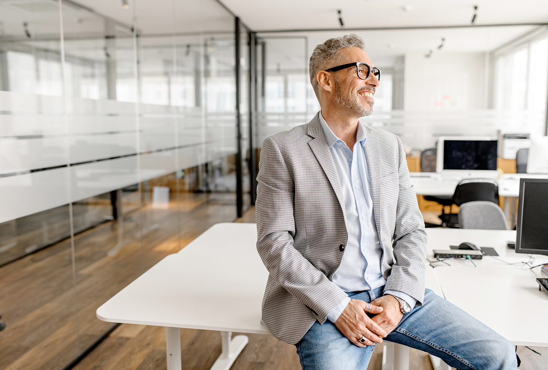 Middle-aged man in glasses and a plaid blazer, smiling while sitting on a desk in a modern, bright office with glass walls and computers.