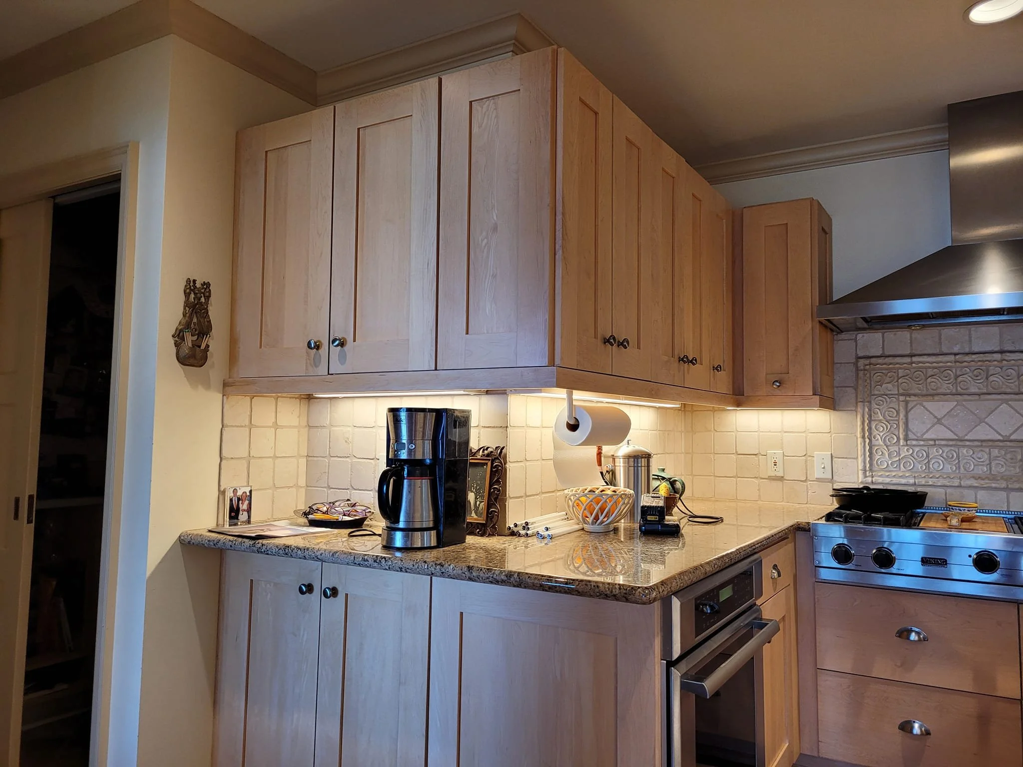 A well-lit kitchen counter with a coffee maker, a bowl of fruit, and other kitchen items. The counter is made of granite and has light wood cabinets below. Above the counter are light wood cabinets, and a stainless steel range hood is visible to the right. A roll of paper towels hangs from under the cabinets.