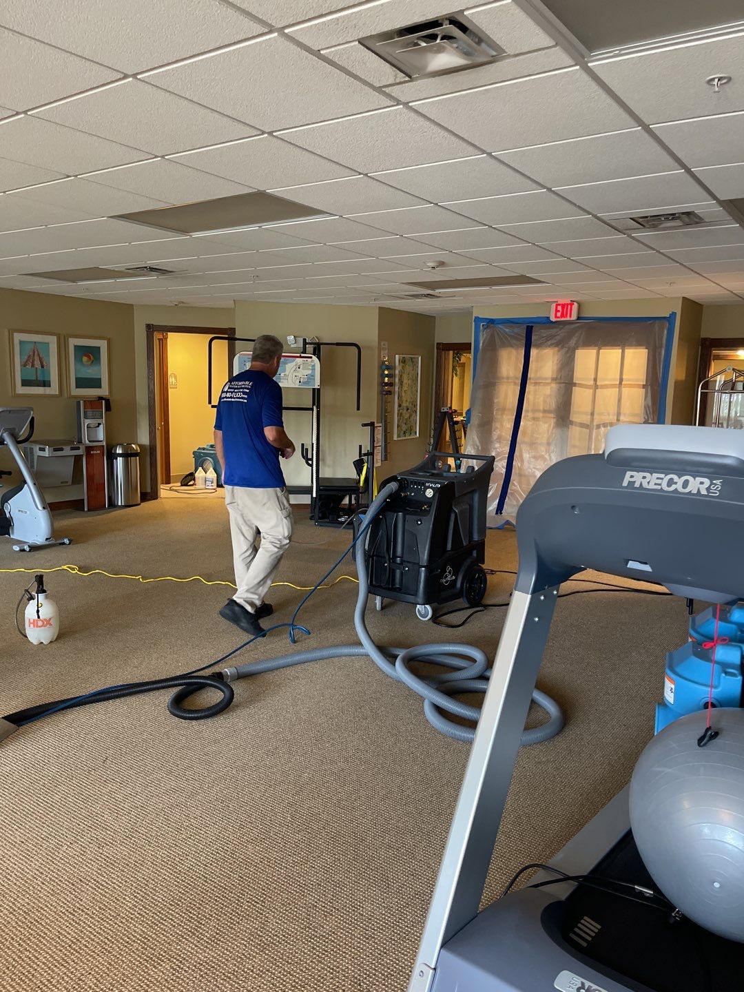 A man in a blue t-shirt and khaki pants is working in a room with exercise equipment and water damage remediation equipment. The room appears to be a fitness center or multipurpose room within a facility. There are treadmills, an exercise bike, and weights. A large, black extraction machine is connected to hoses, and a blue water extraction unit is also visible. A section of the room near the doors is covered with plastic sheeting, suggesting a contained work area for repairs or cleaning. The ceiling has acoustic tiles, and there is an exit sign illuminated above the doors.