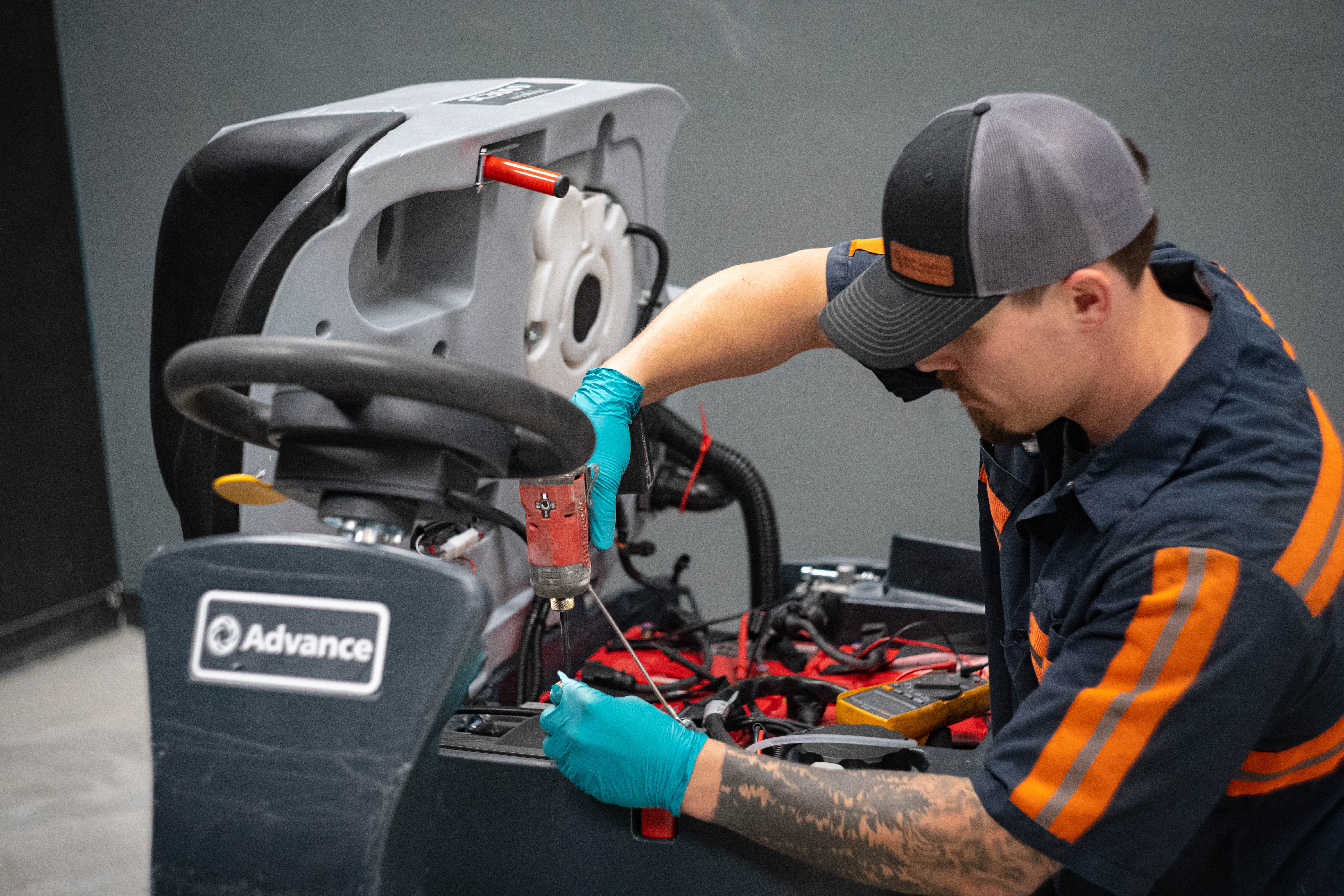 A professional technician with a forearm tattoo works on an Advance SC3000 ride-on floor scrubber. The technician is wearing teal nitrile gloves and using a cordless impact drill to perform precision electrical or internal component maintenance underneath the steering column assembly. The machine's gray body panel is tilted up, exposing internal wiring networks and the battery compartment.