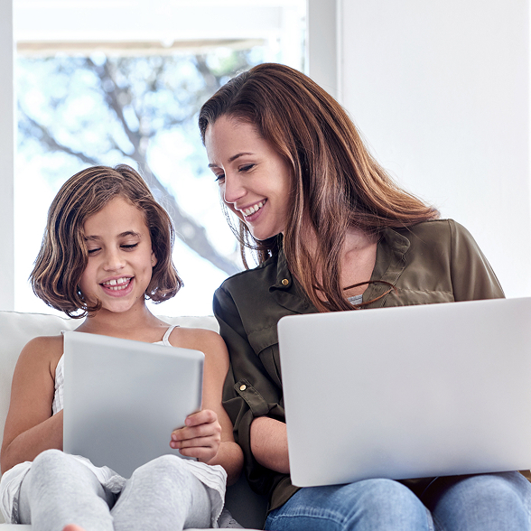 A woman and child sit on a white sofa, smiling and using a laptop and tablet. The room is bright, conveying a warm and happy atmosphere.