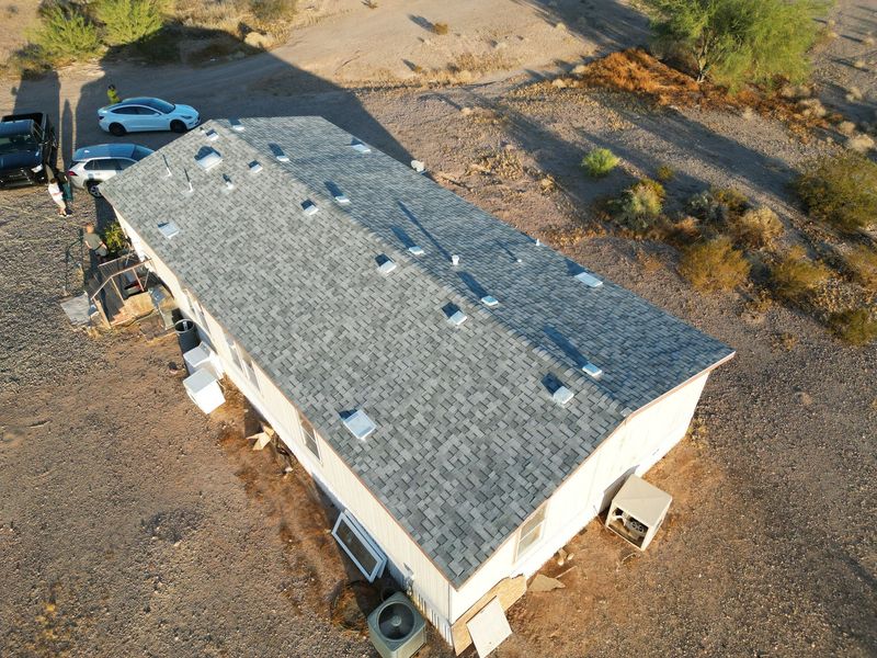 A high-angle aerial drone view displays a completed roofing project on a long, rectangular home. The new gray asphalt shingle roof features multiple white vents and sits within a desert landscape.