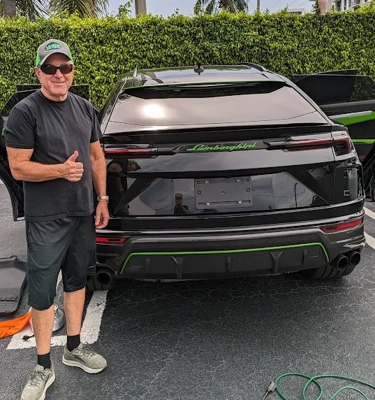 A smiling man in a black t-shirt, black shorts, and a green and gray baseball cap gives a thumbs up next to the rear of a black Lamborghini Urus. The car has green accents on its rear diffuser and the "Lamborghini" lettering on the tailgate is also green. The man is wearing sunglasses and athletic shoes. The car is parked in an outdoor lot with a manicured green hedge in the background and asphalt pavement.