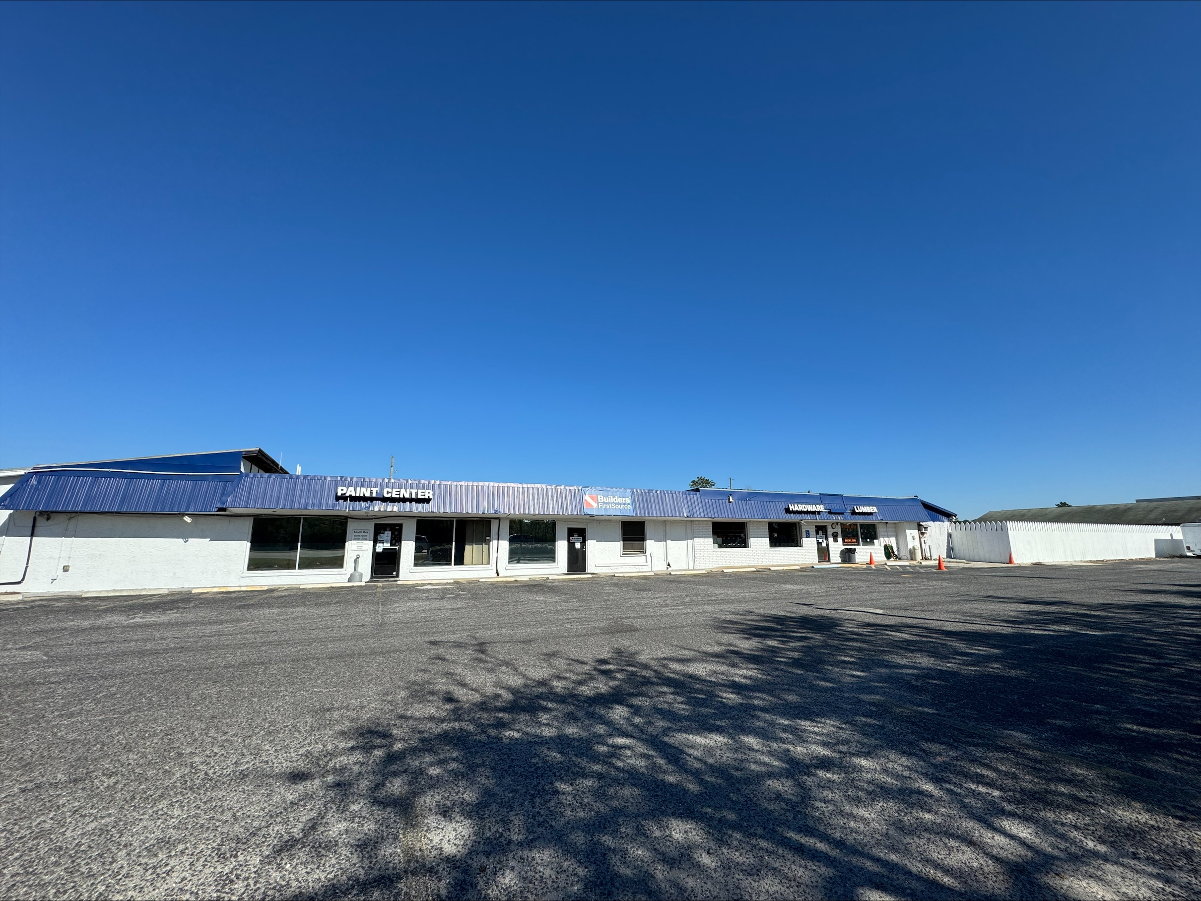 Wide-angle view of the Builders FirstSource Lumber Yard and Parking Lot in Santa Rosa Beach, Florida, highlighting the building's blue awning and ample parking space under a clear blue sky.