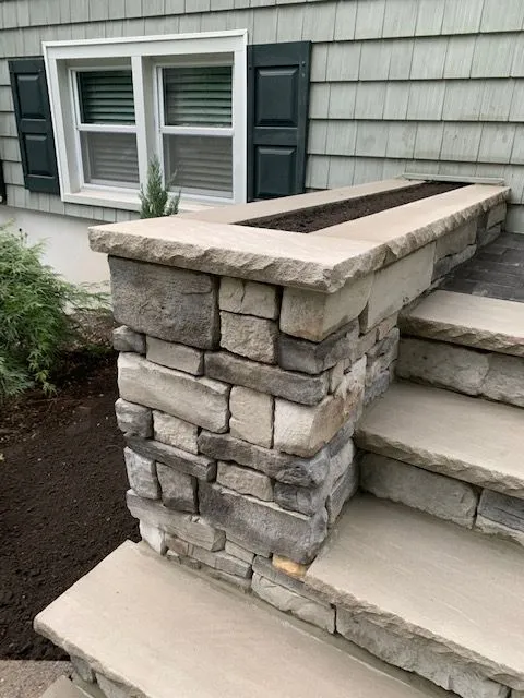 A stone-clad pillar with a planter box on top sits next to concrete steps, leading up to the exterior of a house with gray clapboard siding and dark green shutters. The pillar is constructed from various shades of gray and beige stacked stones. A small evergreen plant peeks out from the planter box. The steps are made of smooth, light-colored concrete.