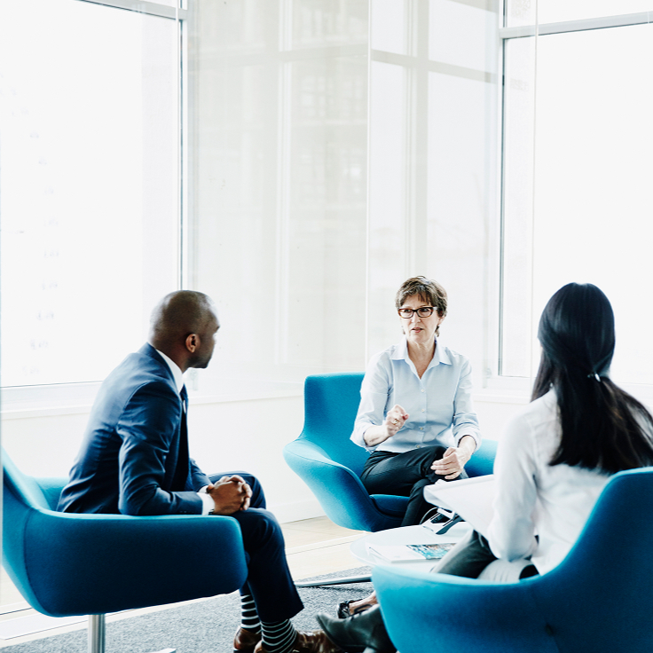 A group of four professionals sits in blue chairs in a modern office with large windows. They are engaged in a serious discussion, fostering a collaborative atmosphere.