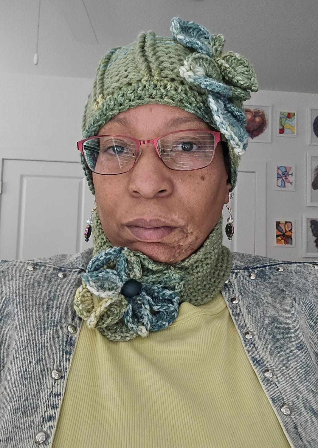 A close-up portrait of a woman wearing a crocheted hat and scarf with floral embellishments. She has on pink-rimmed glasses and a denim jacket over a light yellow ribbed shirt. Several framed pictures are visible on the wall behind her.