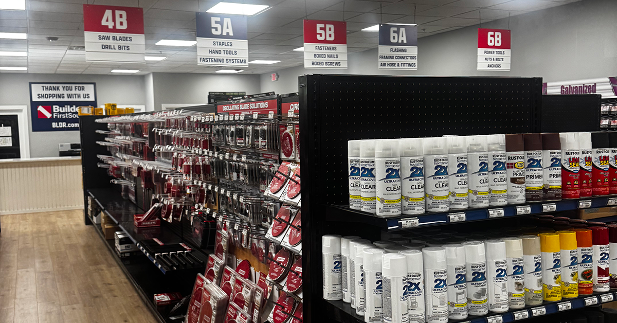 Builders FirstSource store aisle with shelves of Rust-Oleum spray paint in white, black, green, yellow, red and other colors. A neighboring aisle displays packaged saw blades, drill bits, fasteners and hand tools. Overhead signs identify sections 4B through 6B, including saw blades, drill bits, staples, fasteners, boxed nails, power tools and anchors.