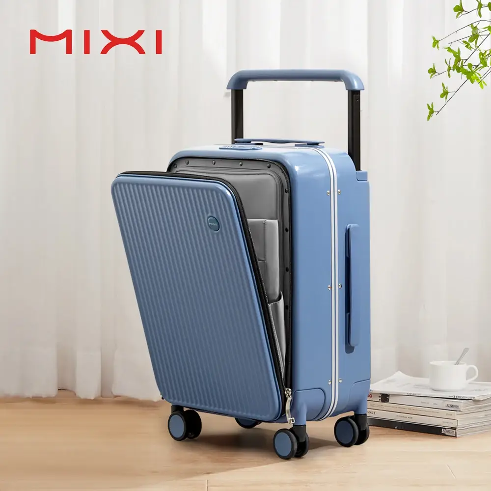 A blue suitcase is open with a laptop inside. The suitcase has a retractable handle and wheels. It is placed on a wooden floor in front of a white curtain. The brand name 'MIXI' is visible in red text at the top left of the image. Several books and a white cup are stacked to the right of the suitcase.