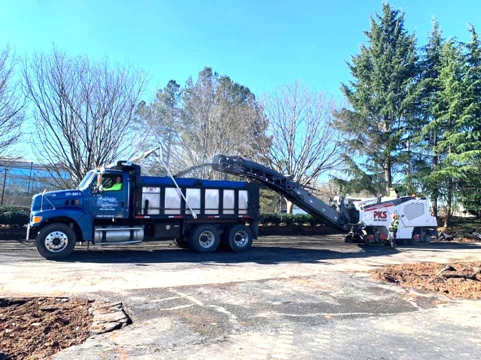 A dump truck is connected to a large milling machine removing asphalt from a road surface. Workers operate the equipment while debris moves up the conveyor into the truck.