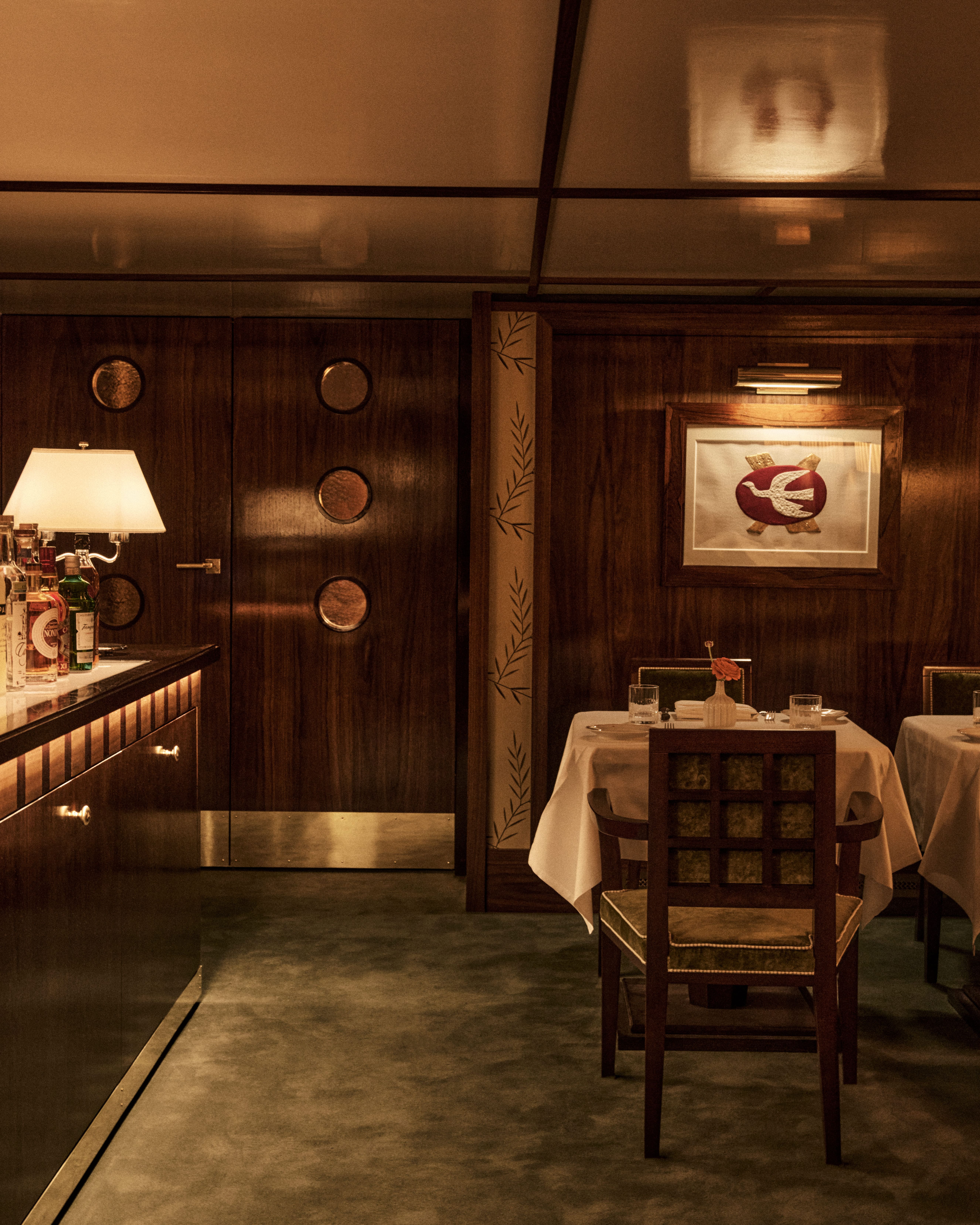 Intimate dining area with dark wood paneling, a small table set with white linens and green-upholstered chair, a softly lit bar counter to the left, and framed nautical-style artwork creating a warm, classic ambiance.