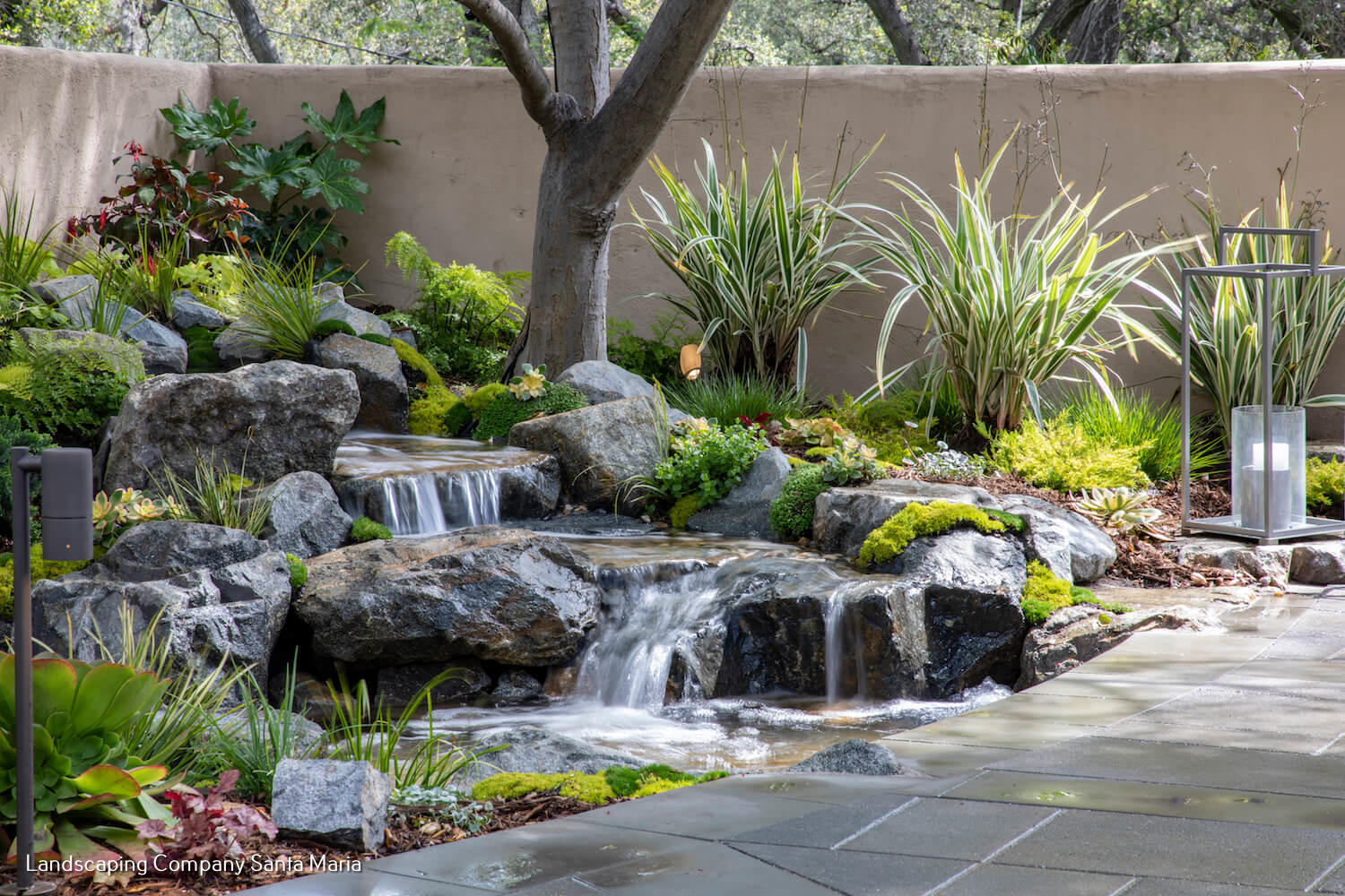 Lush backyard garden with a tiered rock waterfall, pond, ornamental grasses, greenery, and a paved patio creating a tranquil retreat.