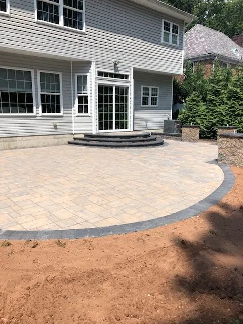 A newly paved patio with tan interlocking pavers is installed next to a grey house with white trim. A set of curved stone steps leads from the patio to a sliding glass door on the back of the house. The patio is bordered by a darker grey paver, and the surrounding ground is covered in reddish-brown dirt, indicating recent landscaping work.