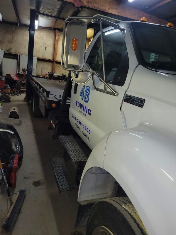 A side perspective photograph captures a white Ford F-750 flatbed tow truck displaying blue "4B Towing" signage, parked indoors within an active service shop surrounded by various vehicle repair equipment.