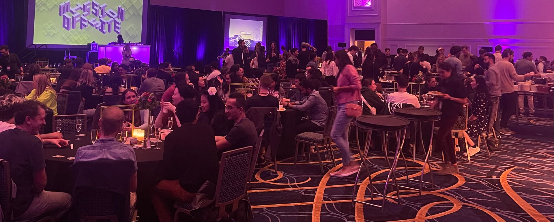 A lively social event with people dining, networking, and enjoying entertainment. A stage with "MISSION OFFSITE" is displayed in the background.